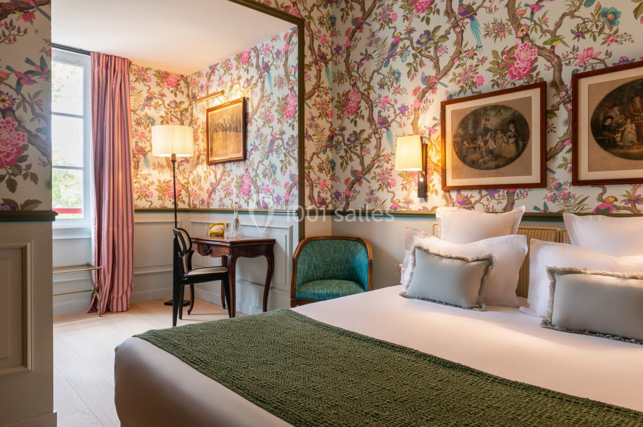 Chambre lumineuse avec lit double, fauteuil, bureau en bois, lampes et murs décorés de papier peint fleuri.