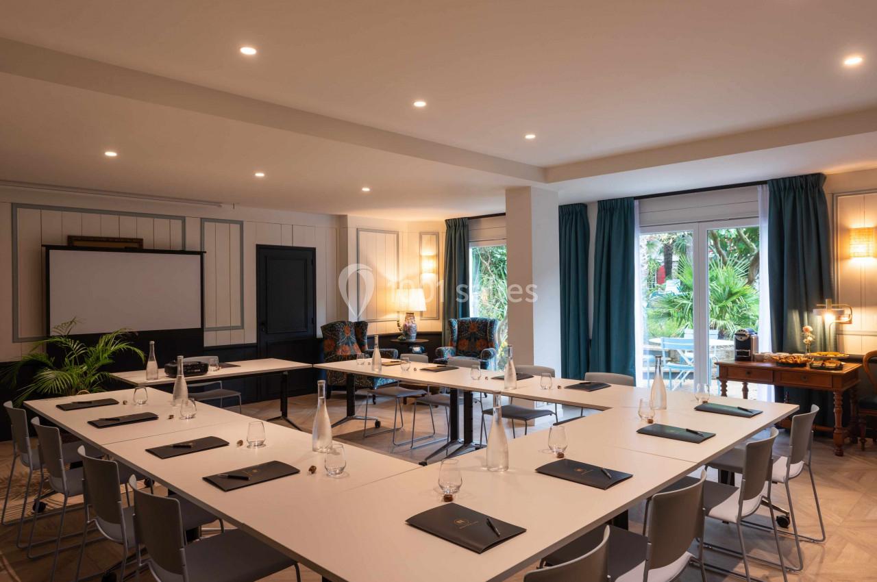 Salle de réunion lumineuse avec tables disposées en U, chaises grises, écran de projection et vue sur un jardin.