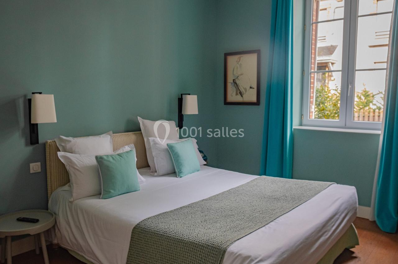 Chambre lumineuse avec lit double, coussins assortis, murs bleu clair, rideaux turquoise et fenêtre donnant sur l'extérieur.