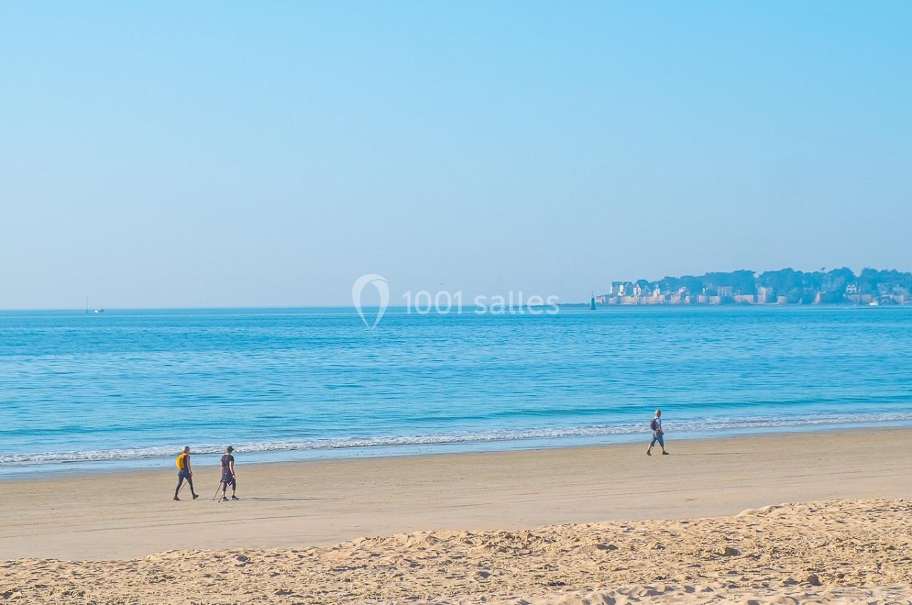Trois personnes marchant sur une plage de sable fin avec une mer calme et un ciel bleu en arrière-plan.