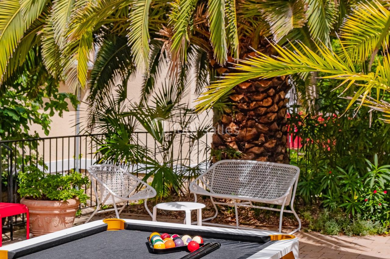 Table de billard extérieure blanche avec accessoires, placée sur une terrasse pavée entourée de palmiers et de mobilier de…