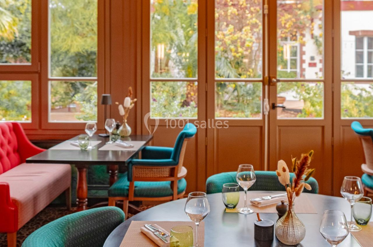 Salle de restaurant lumineuse avec tables dressées, chaises vertes et grandes baies vitrées donnant sur un jardin.