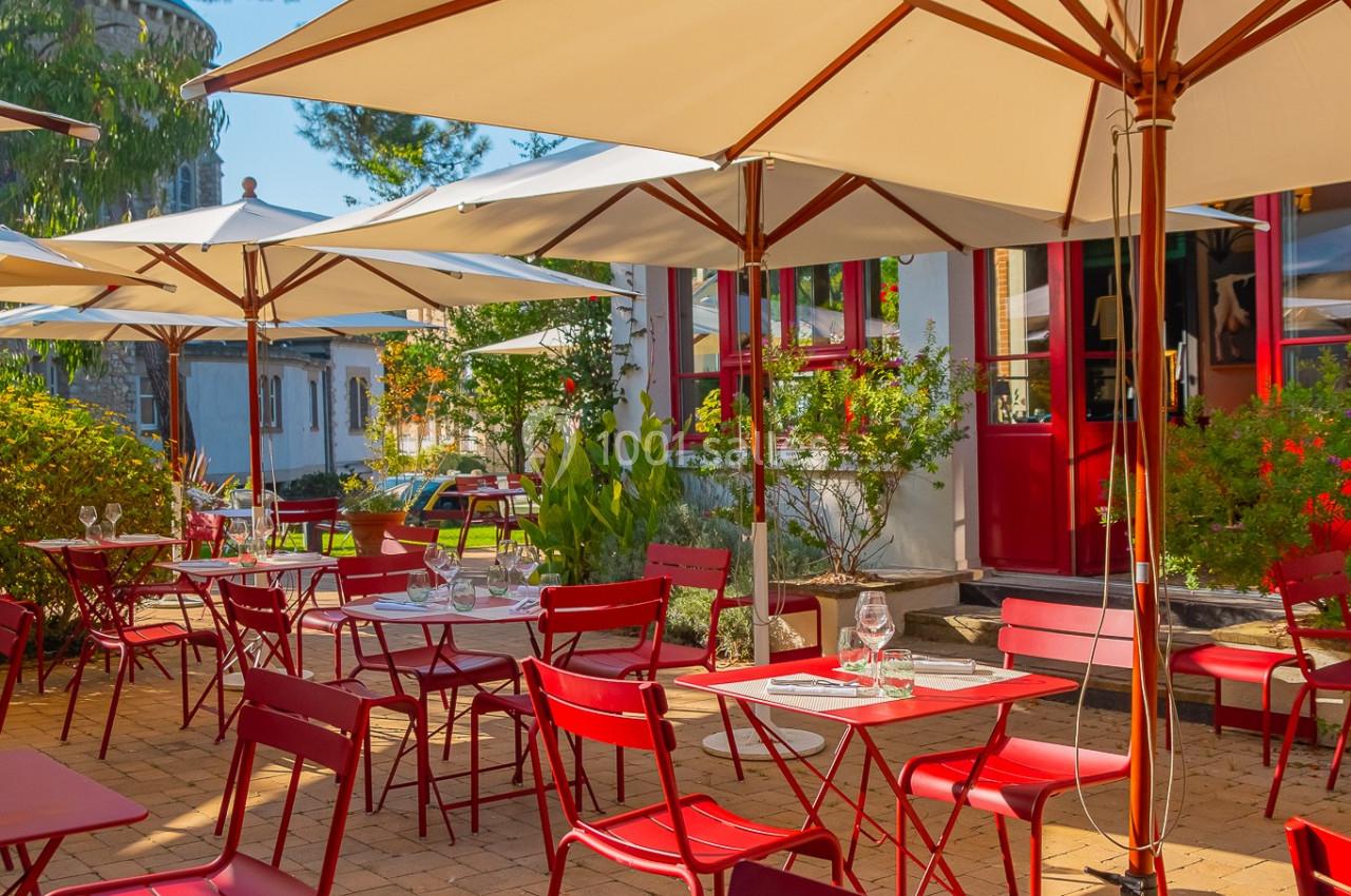 Terrasse ensoleillée avec tables et chaises rouges, entourée de parasols et de végétation, devant un bâtiment.