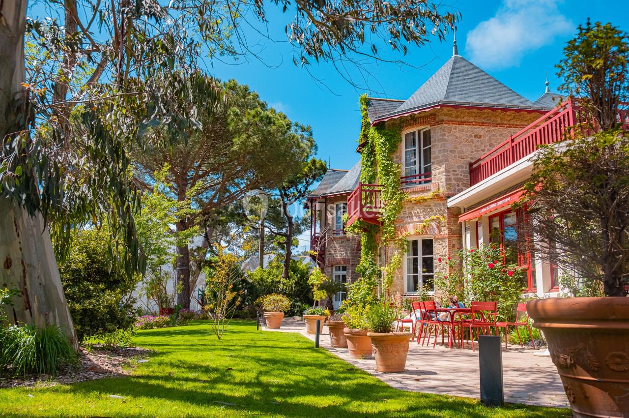 Maison en pierre avec volets rouges entourée d'un jardin verdoyant et ensoleillé, avec terrasse et pots de fleurs.