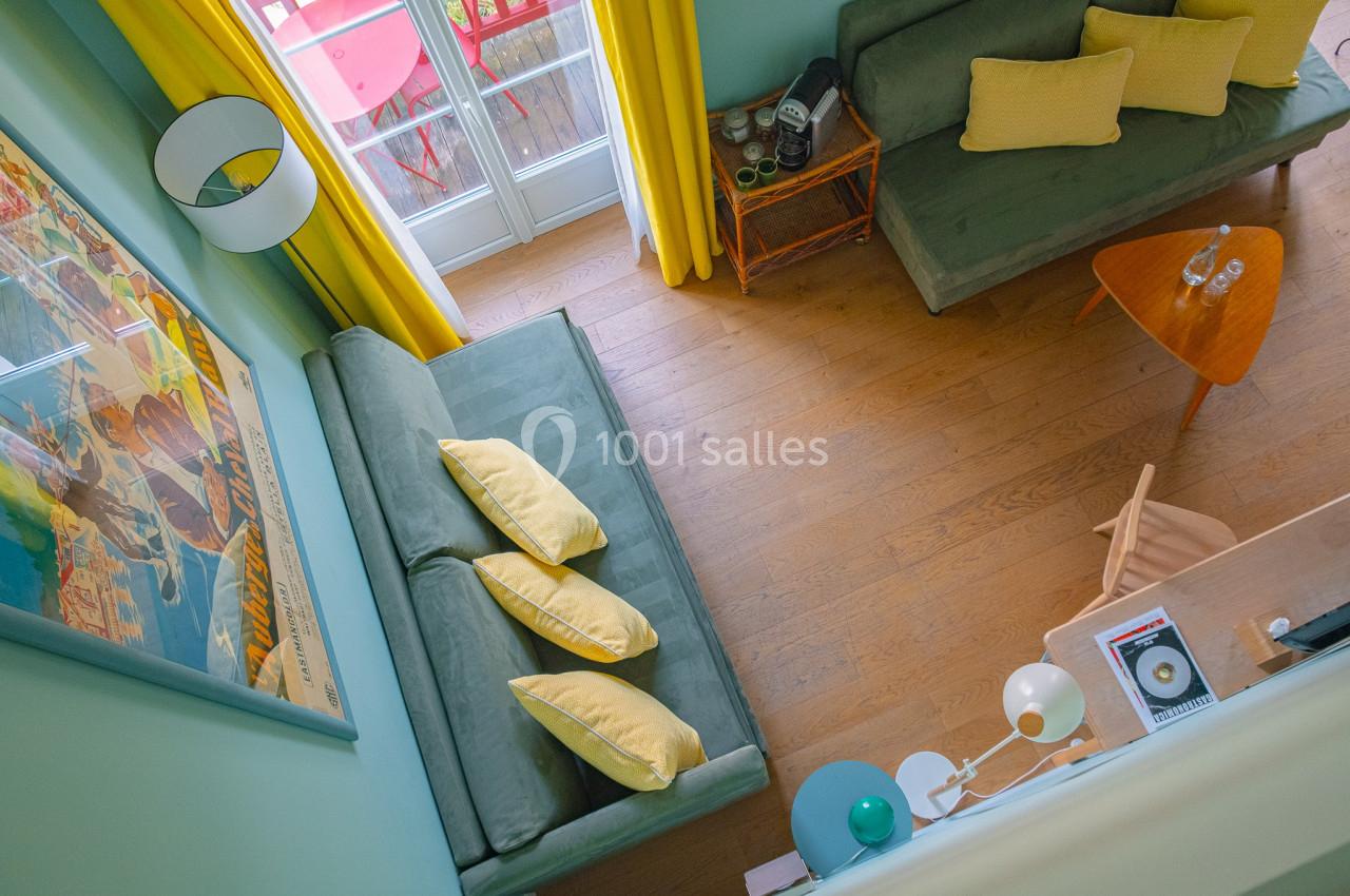 Vue plongeante d'un salon coloré avec canapés verts, coussins jaunes, table basse en bois et porte-fenêtre donnant sur un…