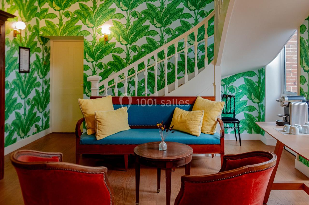 Salon coloré avec canapé bleu, coussins jaunes, fauteuils rouges, table basse ronde et papier peint à motifs de feuilles…