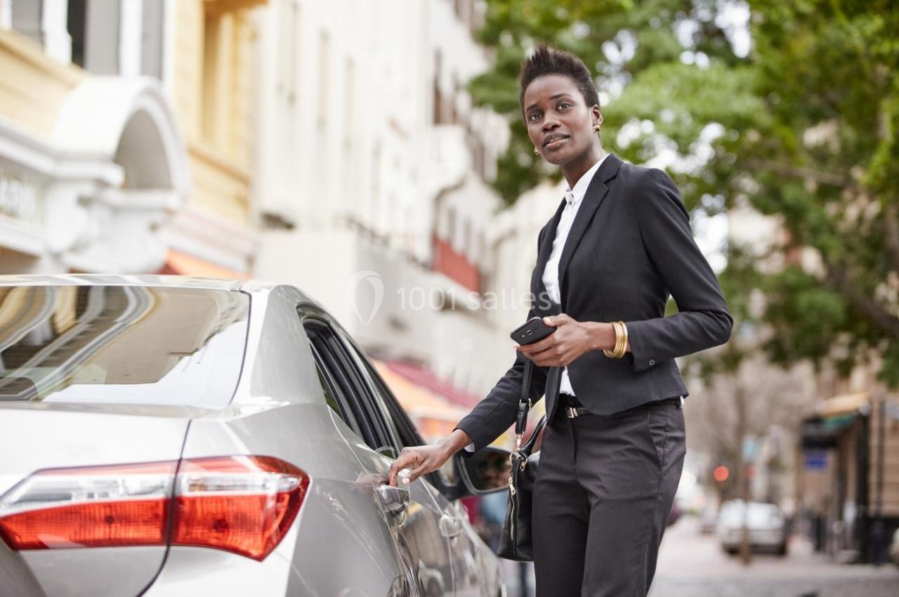 Une femme en tenue professionnelle ouvre la portière d'une voiture argentée dans une rue bordée d'arbres.