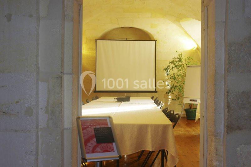Salle de réunion avec table rectangulaire, chaises, écran de projection, tableau blanc et plante en arrière-plan.