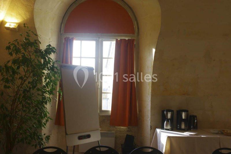 Salle lumineuse avec un tableau de conférence, des rideaux orange, une plante et une table avec des cafetières.