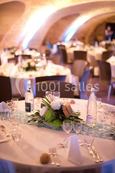 Salle de réception avec tables rondes décorées de centres de table floraux, nappes blanches et bouteilles d'eau.