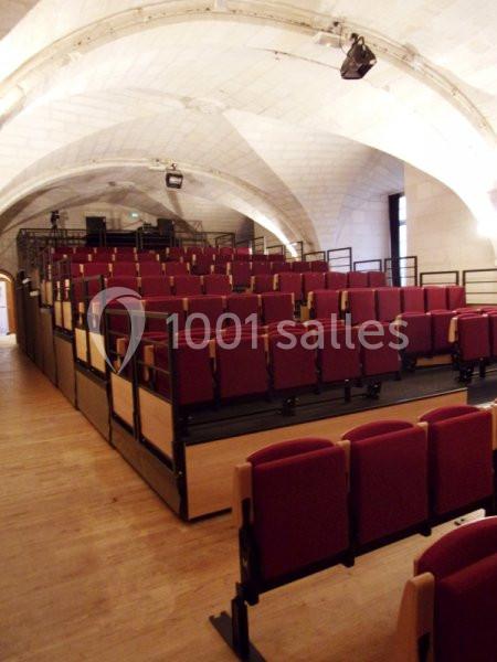Salle avec des rangées de sièges rouges alignés, dans un espace voûté en pierre avec un sol en bois.