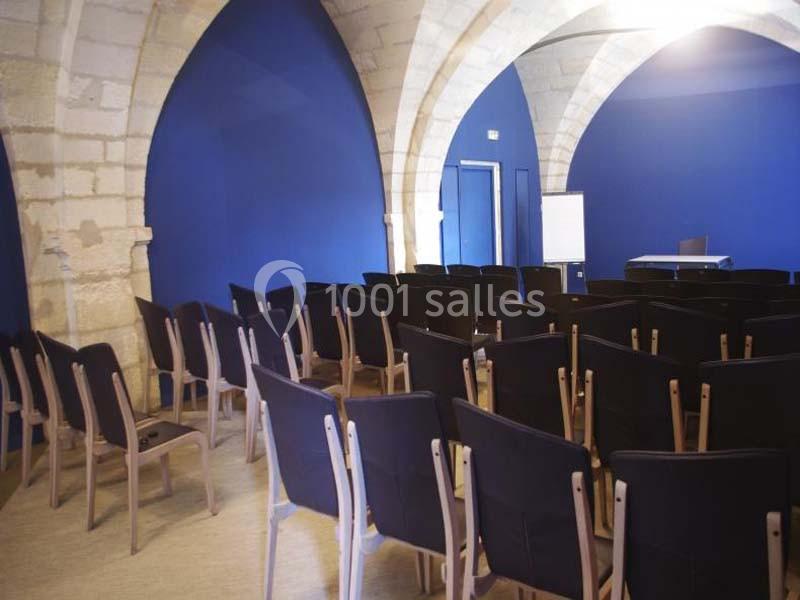 Salle voûtée avec murs bleus, rangées de chaises noires alignées face à un tableau blanc et une table.