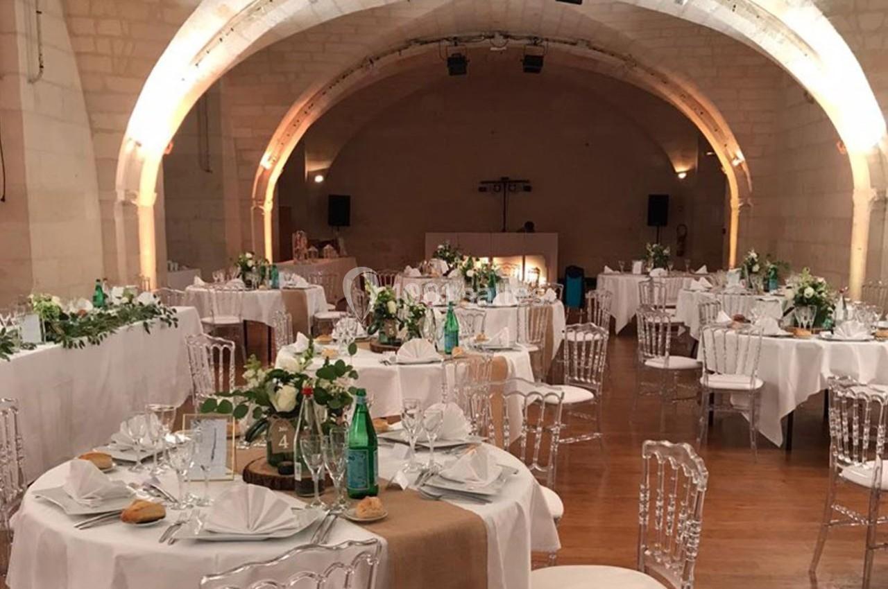 Salle voûtée décorée pour un événement, avec des tables rondes dressées, nappes blanches et chaises transparentes.