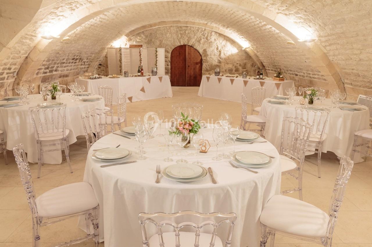 Salle voûtée décorée pour un événement, avec tables rondes dressées, chaises transparentes et éclairage chaleureux.