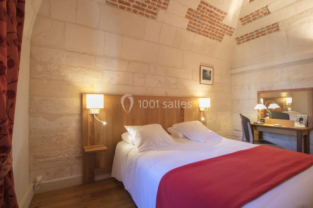 Chambre avec lit double, mur en pierre apparente, bureau en bois, miroir et éclairage chaleureux.