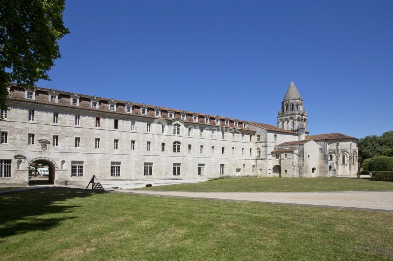 Façade d'un bâtiment historique en pierre avec une tour, entouré d'une pelouse et d'arbres sous un ciel bleu.