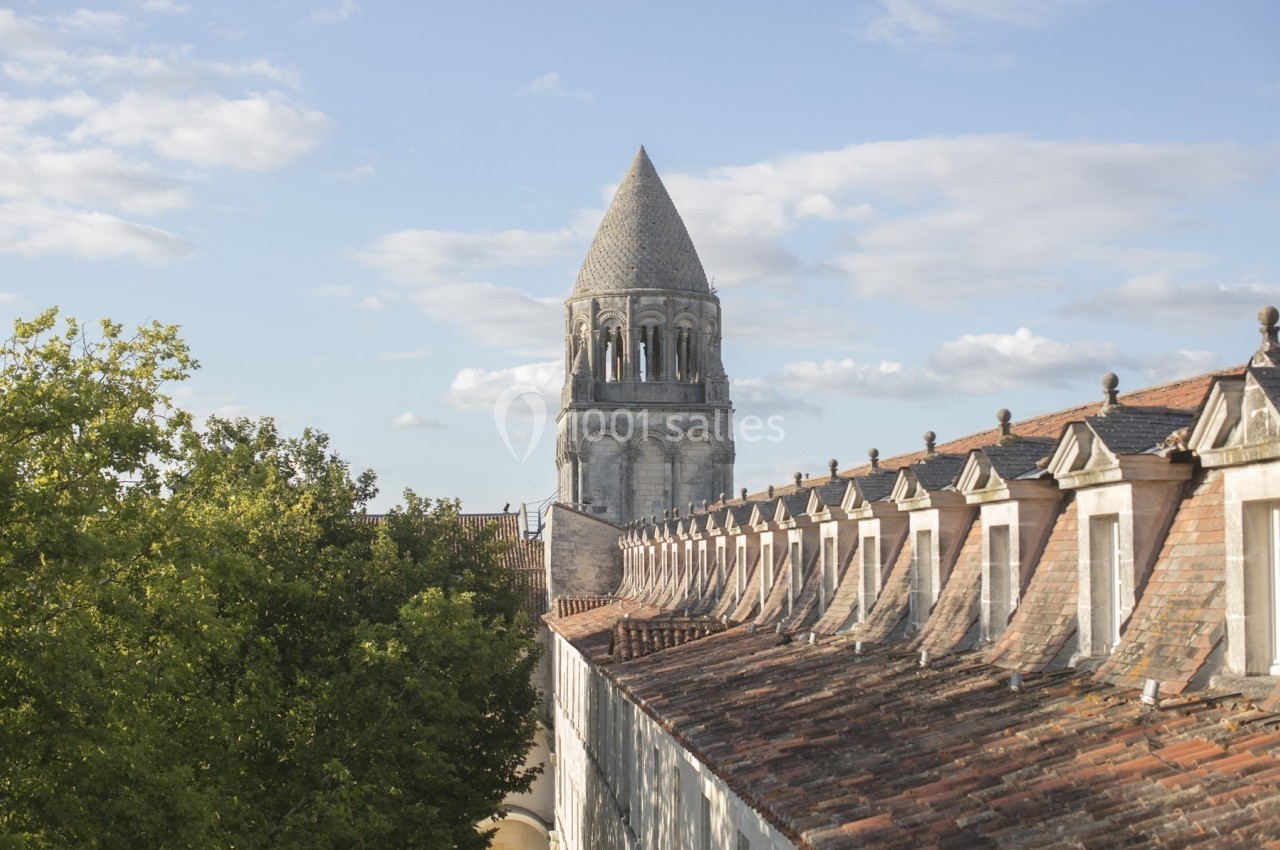 Clocher d'une église romane entouré de toits en tuiles et d'arbres sous un ciel partiellement nuageux.
