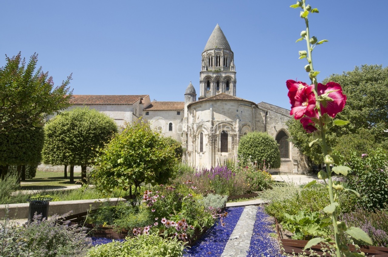 Jardin fleuri avec allée centrale et vue sur une église romane sous un ciel bleu clair.