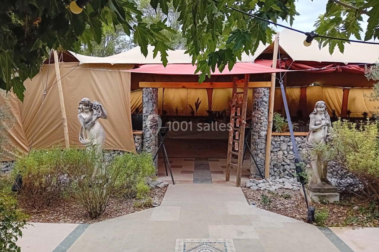 Entrée d'une tente beige entourée de statues décoratives et de végétation, avec un chemin dallé au premier plan.