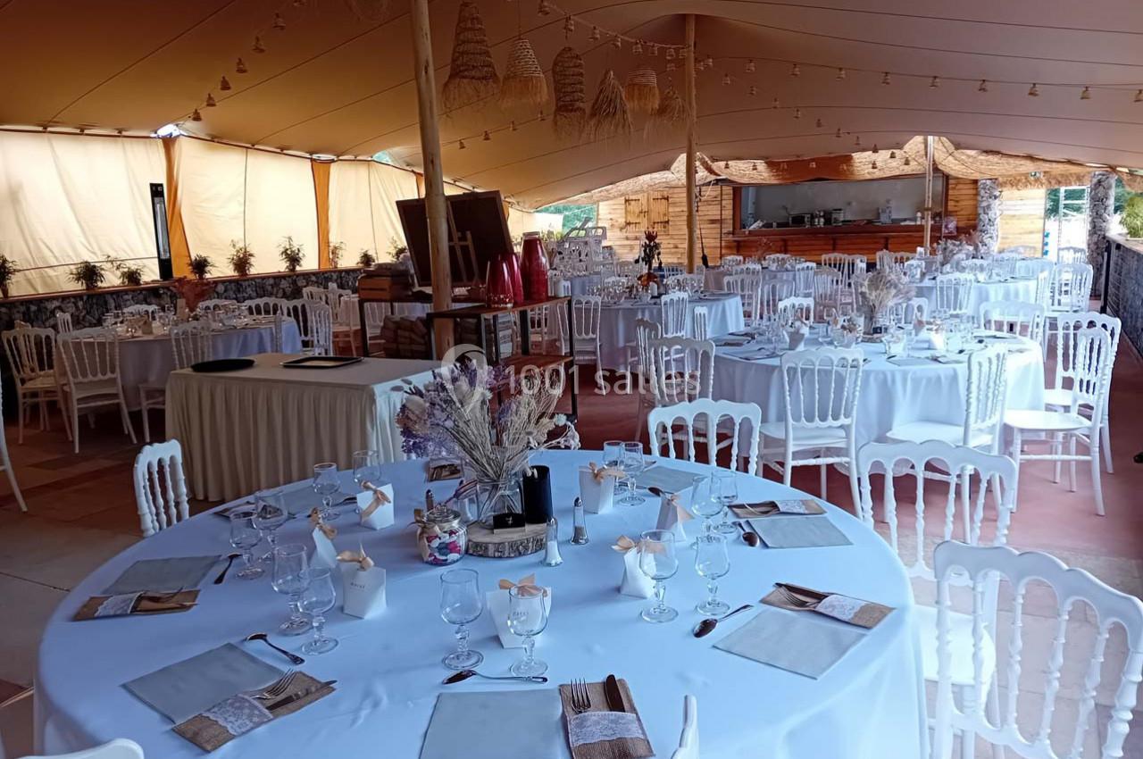 Salle de réception décorée avec des tables rondes dressées, chaises blanches et ambiance lumineuse sous une tente.