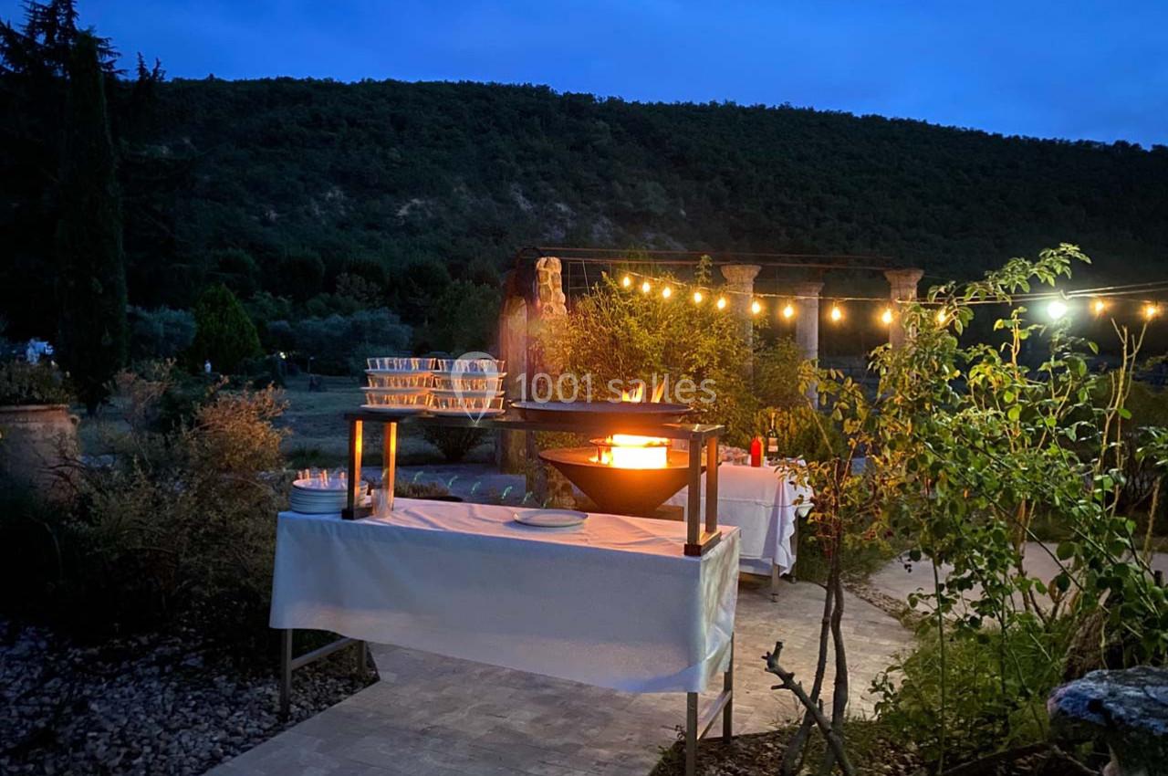Table dressée en extérieur au crépuscule, avec un brasero allumé et des guirlandes lumineuses dans un cadre naturel.