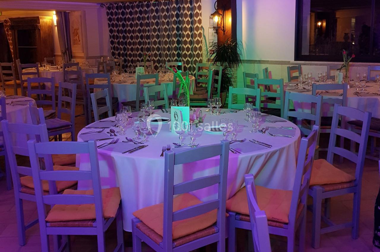 Salle de réception avec tables rondes dressées, nappes blanches, chaises en bois et éclairage coloré.