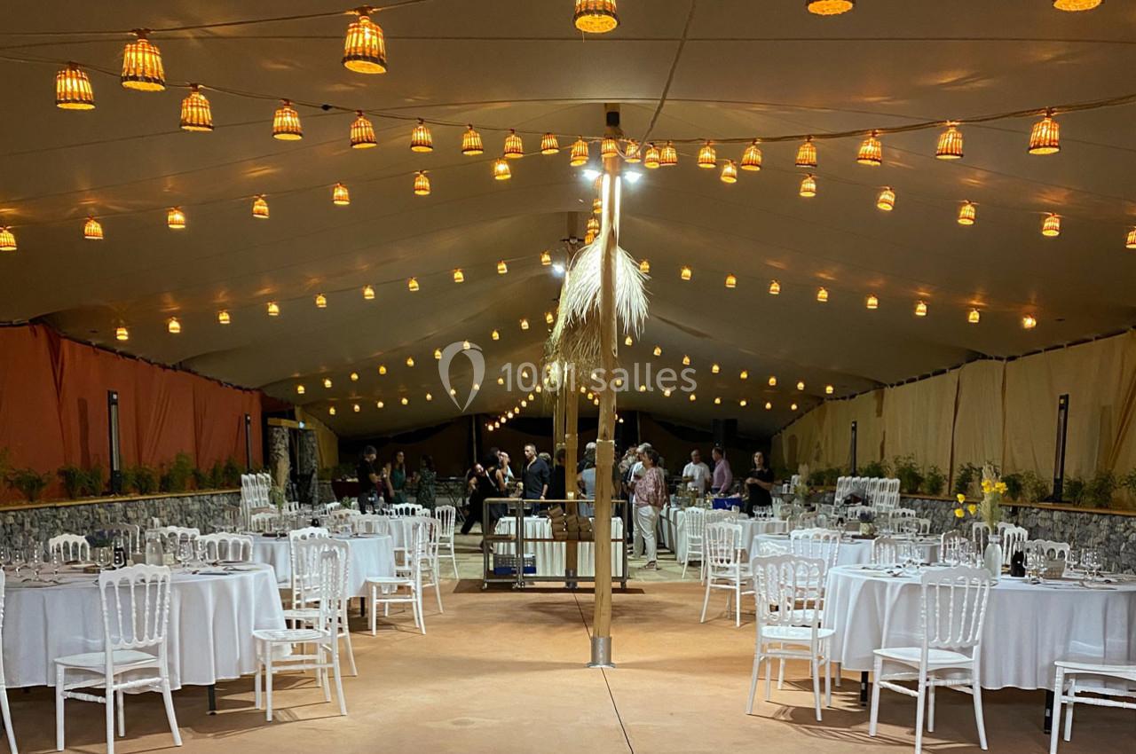 Salle de réception sous une tente éclairée, avec des tables rondes dressées et des invités en arrière-plan.