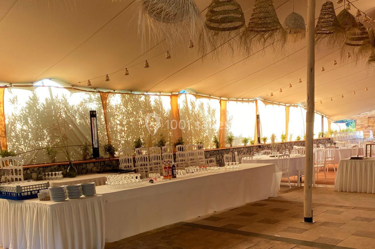 Salle de réception en plein air avec tables dressées, chaises blanches et décorations suspendues sous une tente.