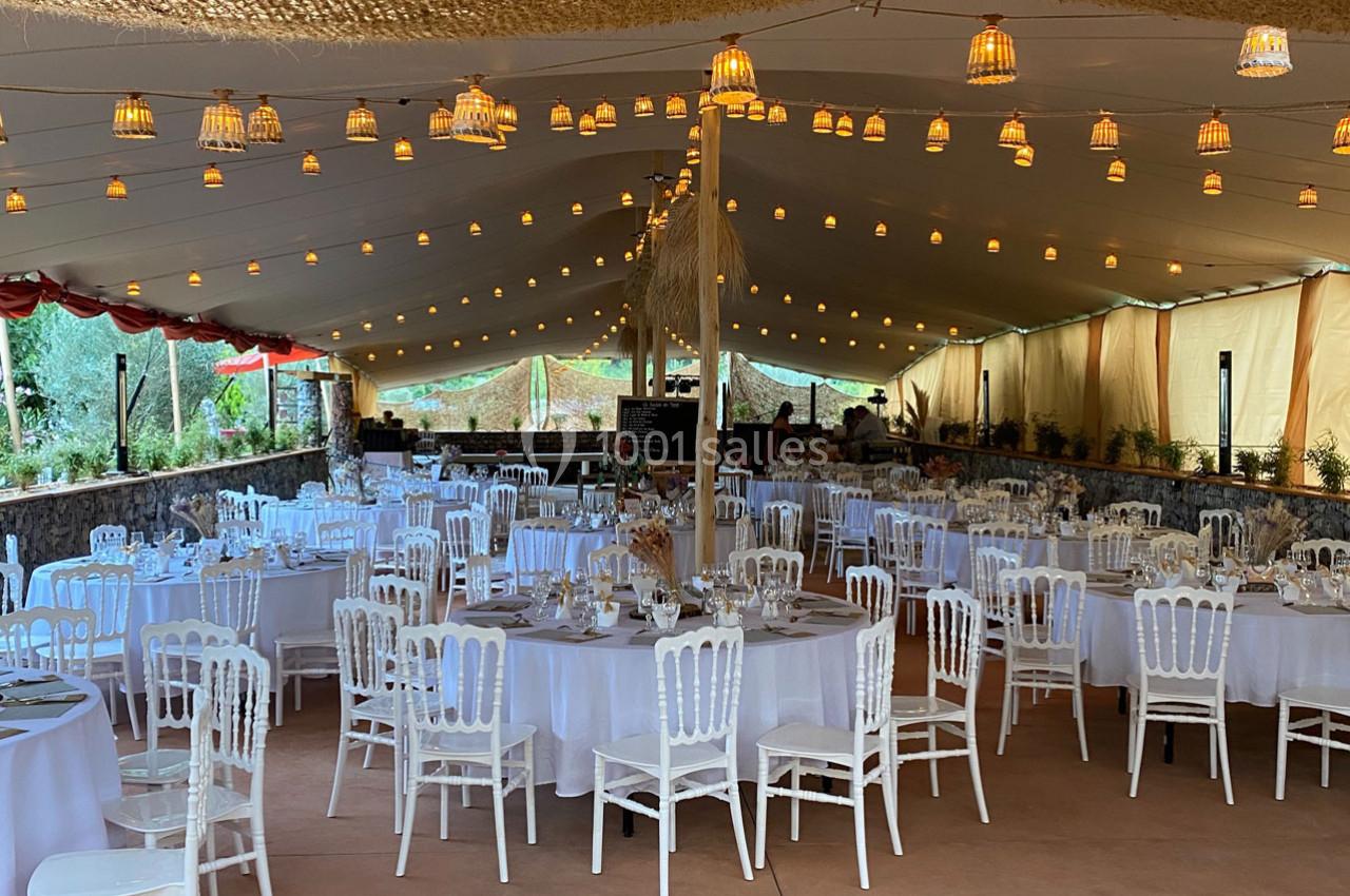 Salle de réception décorée avec des tables rondes dressées, des chaises blanches et des guirlandes lumineuses suspendues.