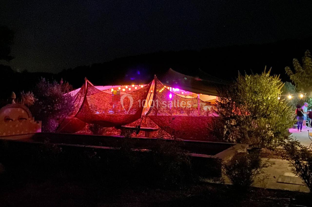 Tente éclairée par des guirlandes lumineuses dans un jardin sombre, entourée de végétation.
