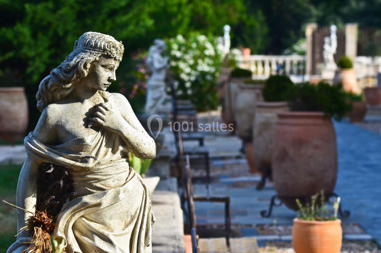 Statue en pierre représentant une femme dans un jardin avec des pots en terre cuite et une allée pavée en arrière-plan.