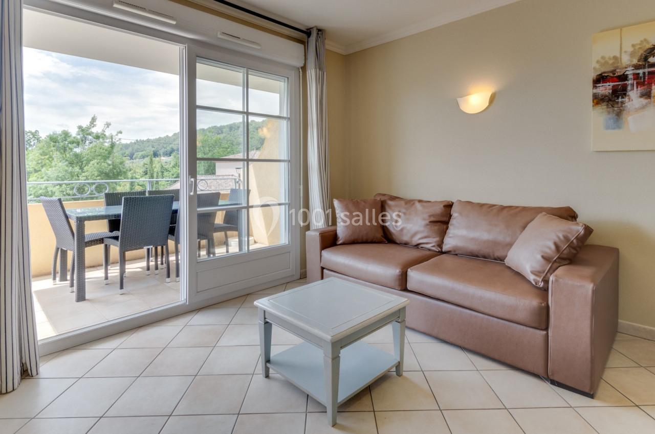 Salon lumineux avec canapé en cuir brun, table basse et accès à un balcon meublé donnant sur un paysage verdoyant.
