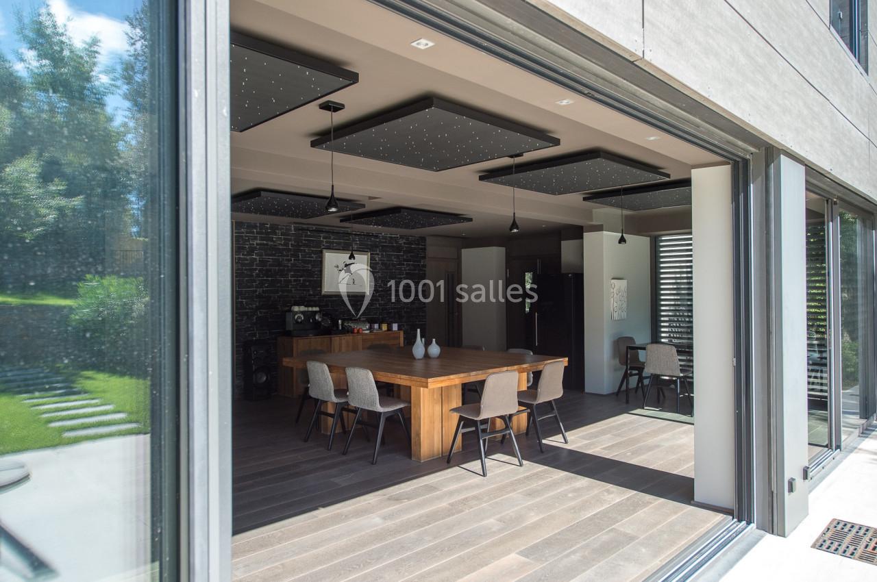 Salle à manger moderne avec une grande table en bois, chaises grises et baies vitrées donnant sur un jardin verdoyant.