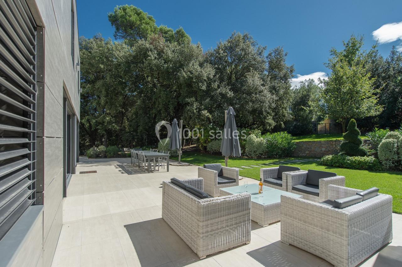 Terrasse avec mobilier en rotin, table basse en verre, parasols et vue sur un jardin arboré par temps ensoleillé.