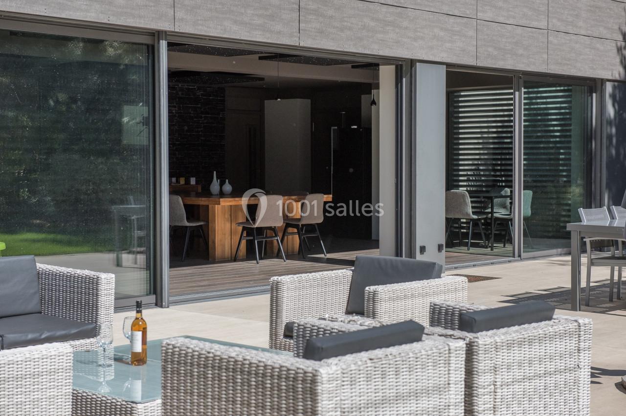 Espace extérieur avec fauteuils en rotin et table basse, donnant sur une salle à manger moderne avec grandes baies vitrées.