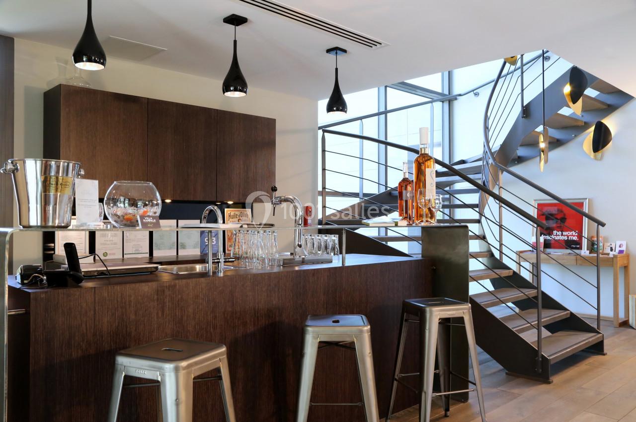 Bar moderne avec comptoir en bois sombre, tabourets métalliques, étagères avec bouteilles et escalier en arrière-plan.