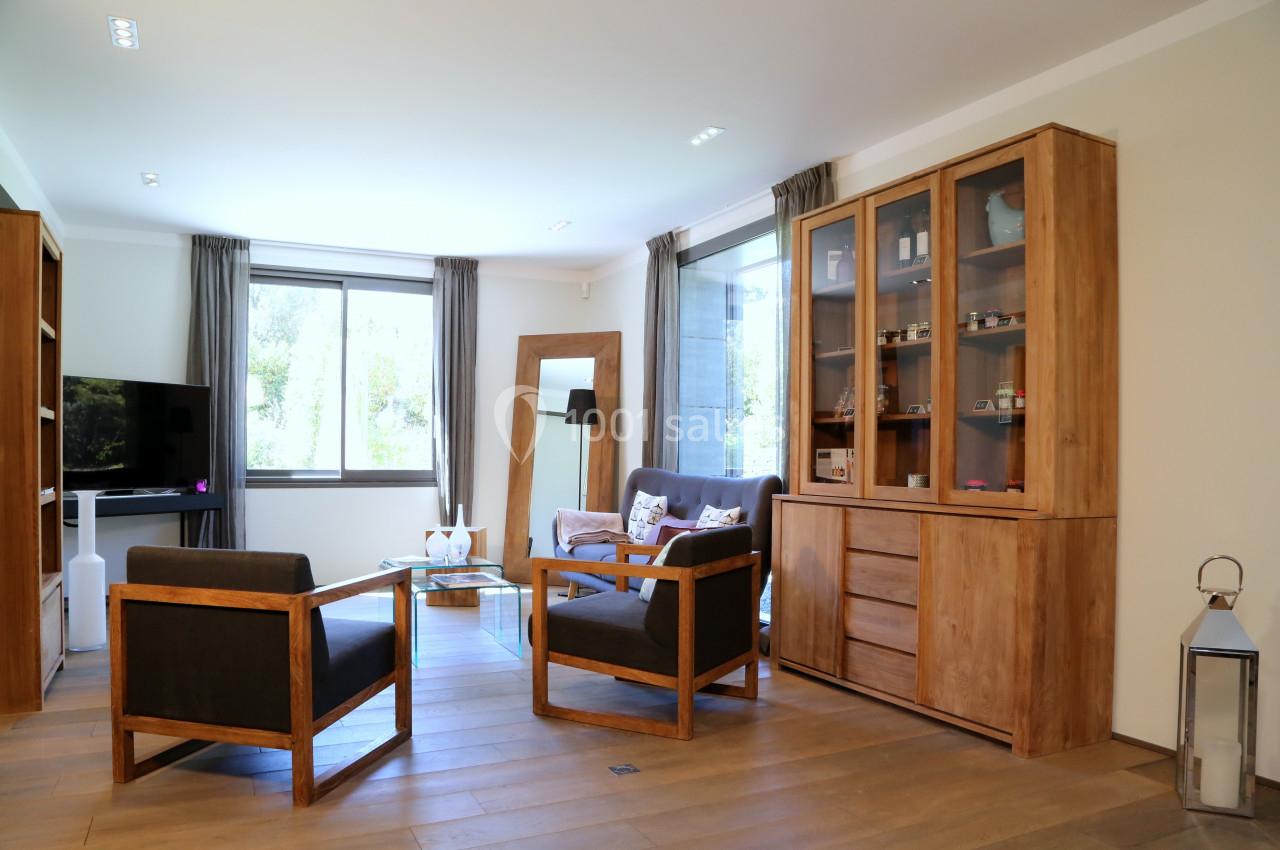 Salon lumineux avec mobilier en bois, fauteuils noirs, grande fenêtre et étagères vitrées contenant des objets décoratifs.