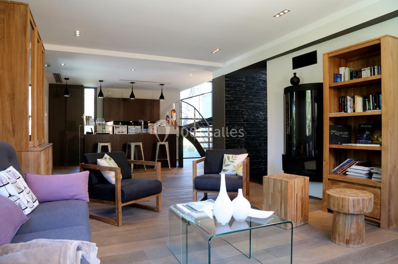 Salon moderne avec mobilier en bois, bibliothèque, poêle noir, et vue sur une cuisine ouverte avec un escalier en arrière…