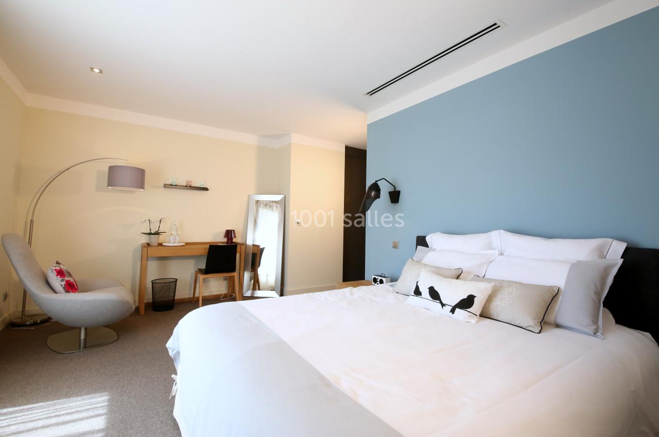 Chambre lumineuse avec lit double, mur bleu, bureau en bois, fauteuil gris et miroir sur pied.