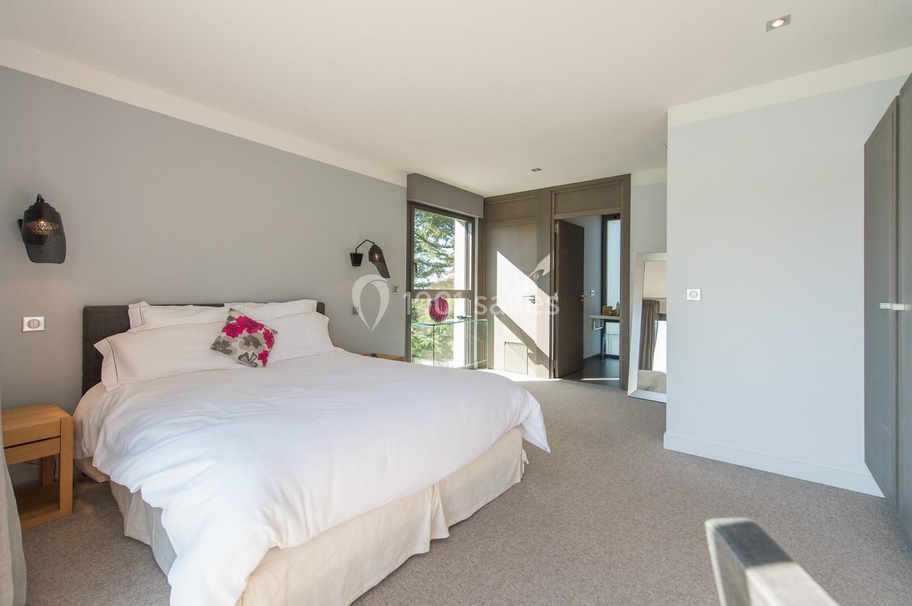 Chambre lumineuse avec lit double, linge blanc, murs gris clair, grandes fenêtres et mobilier en bois moderne.