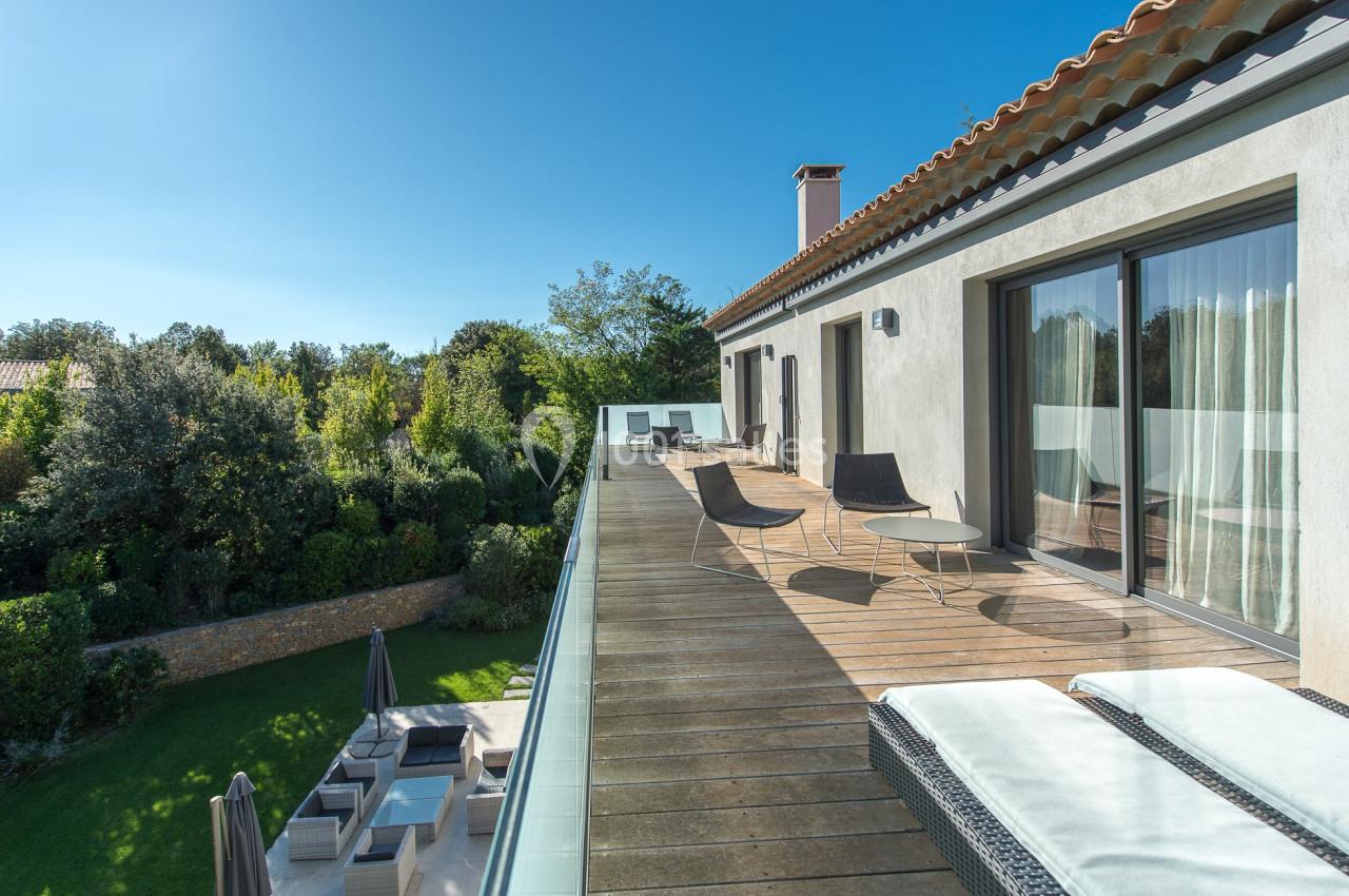 Terrasse en bois avec chaises longues et vue sur un jardin arboré, attenante à une maison moderne sous ciel dégagé.