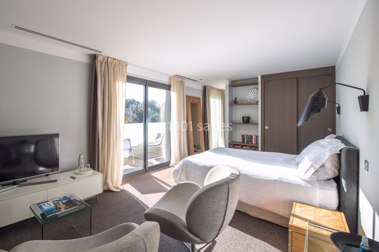 Chambre lumineuse avec lit double, fauteuils, télévision, grande baie vitrée et accès à un balcon.