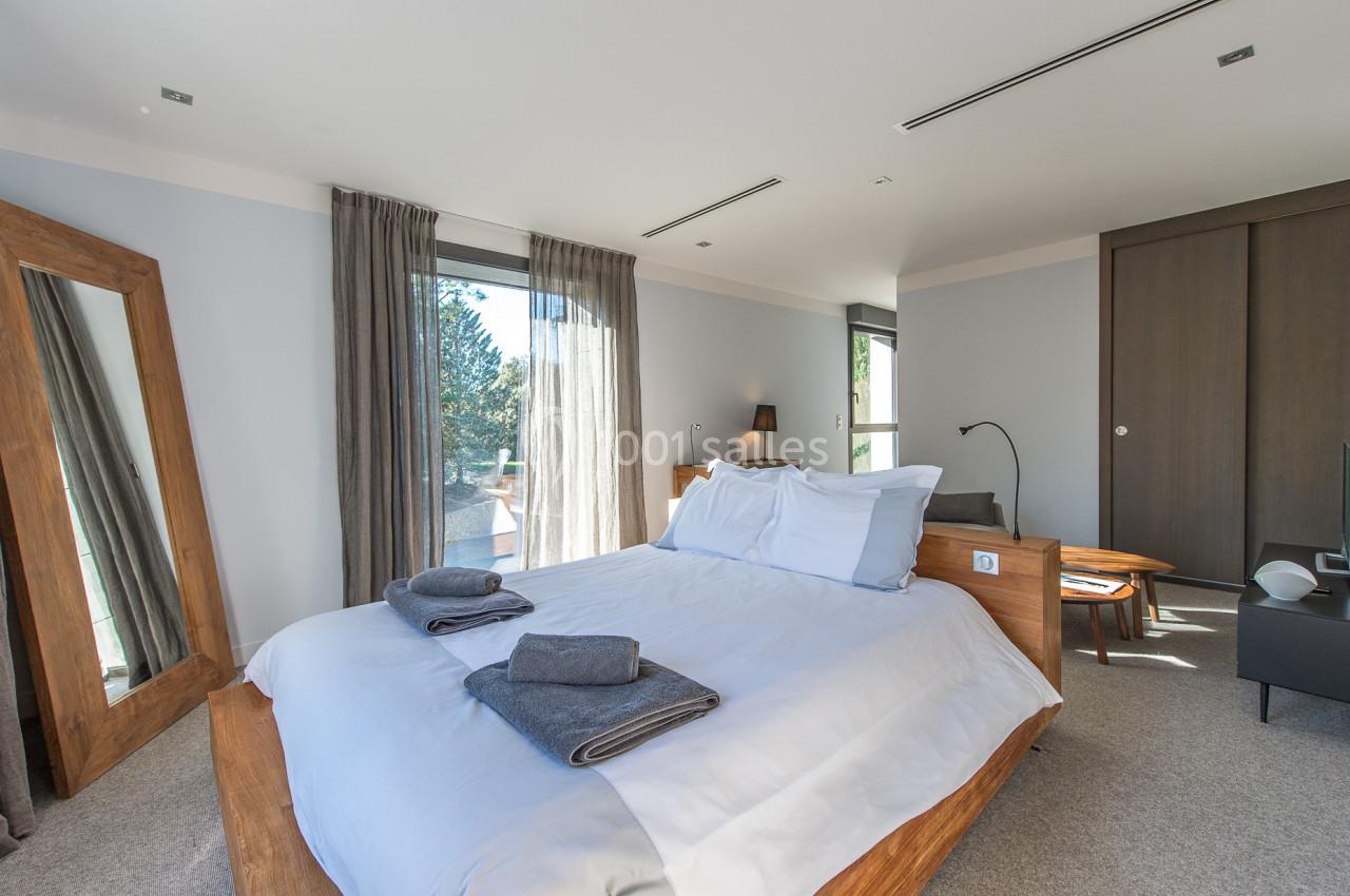 Chambre lumineuse avec lit double, miroir en bois, grandes fenêtres avec rideaux et armoire encastrée.
