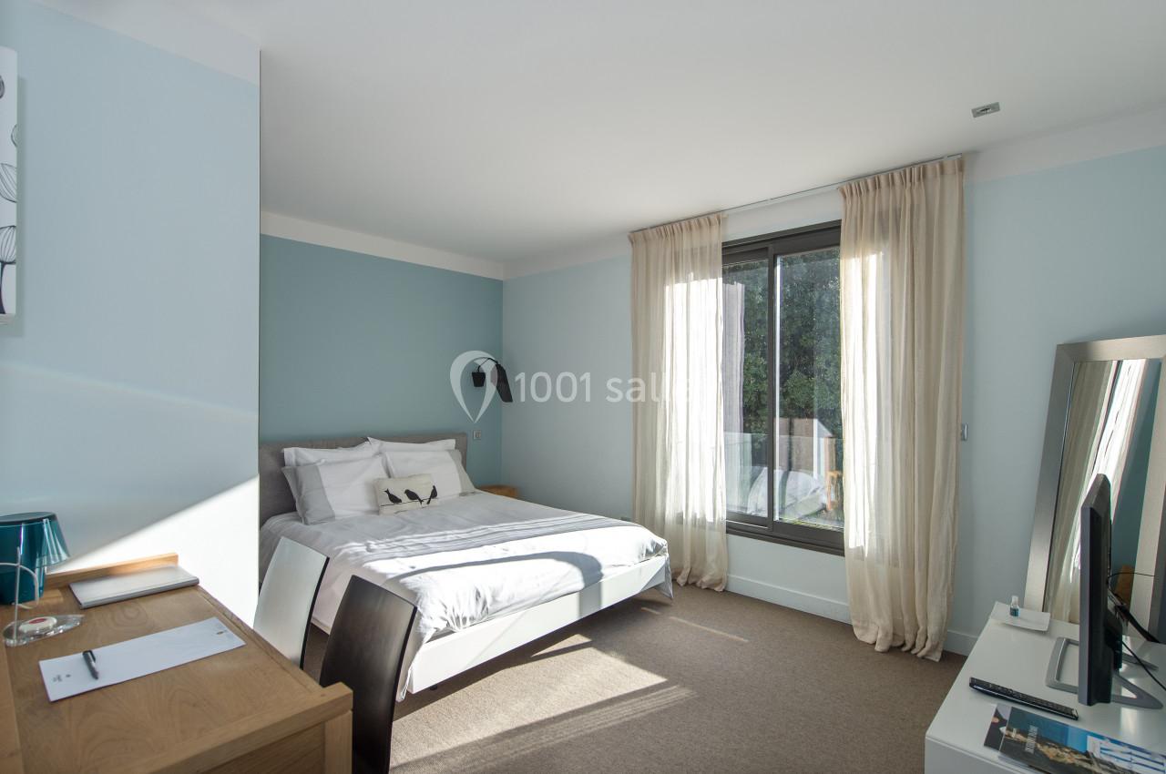 Chambre lumineuse avec lit double, bureau en bois, rideaux beiges et grande fenêtre donnant sur un extérieur verdoyant.