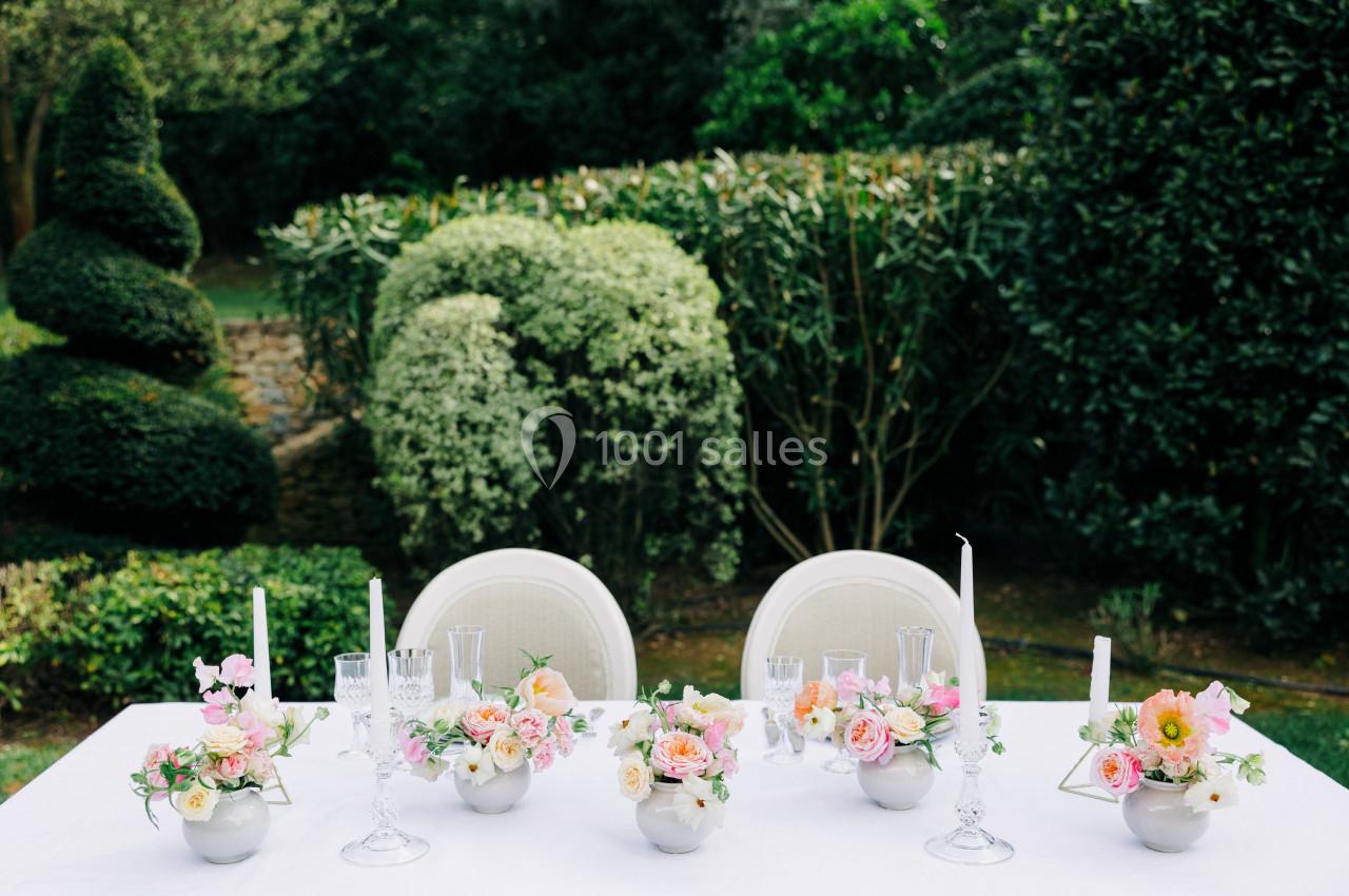 Table décorée avec des fleurs colorées, des bougies blanches et de la vaisselle, placée dans un jardin verdoyant.