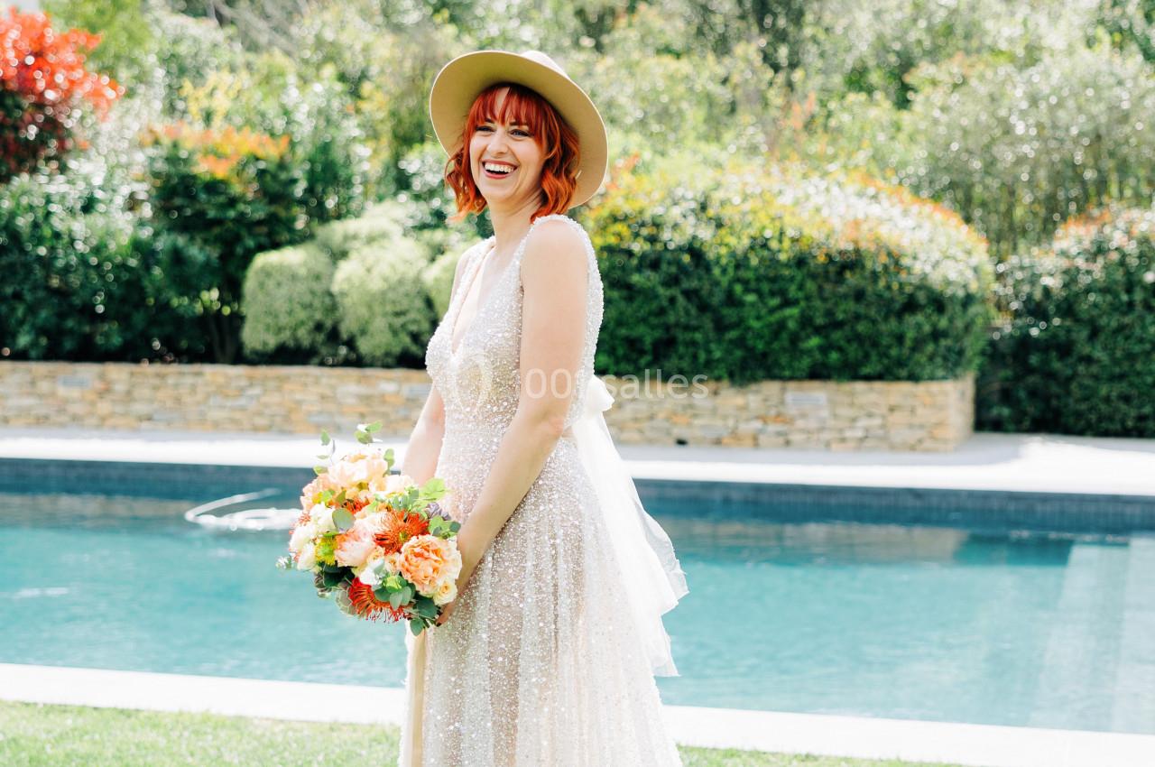 Femme souriante en robe de mariée blanche tenant un bouquet coloré, debout près d'une piscine dans un jardin.