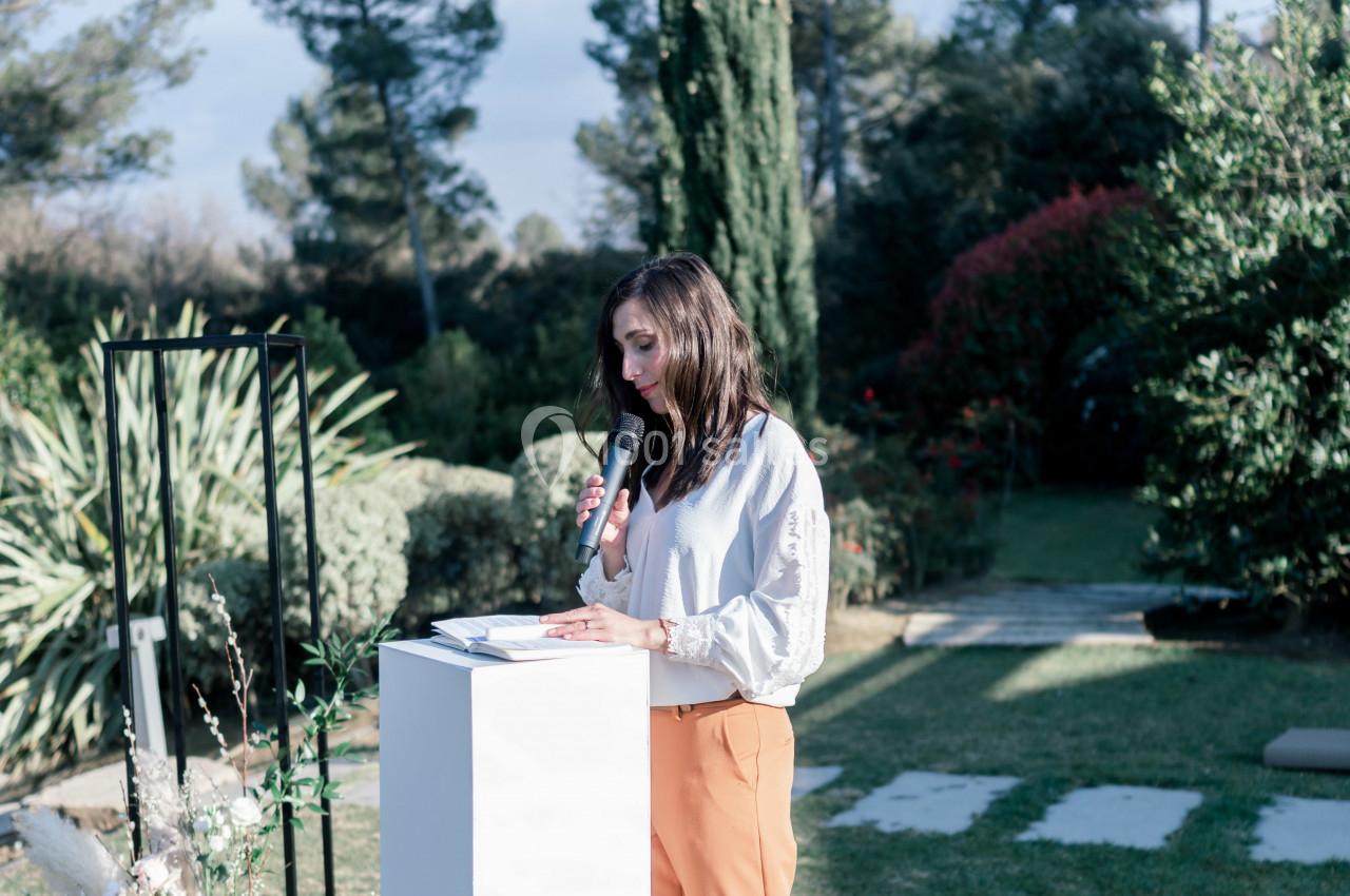 Une femme tenant un micro parle debout derrière un pupitre blanc dans un jardin arboré.