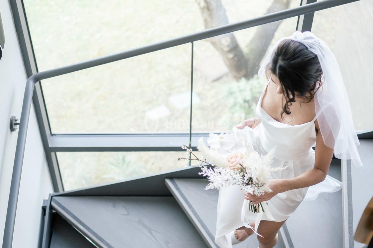 Une mariée en robe blanche et voile monte un escalier en colimaçon, tenant un bouquet de fleurs pastel.