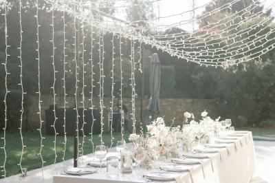 Table dressée pour un dîner en extérieur sous une structure lumineuse avec guirlandes, près d'un bâtiment moderne.