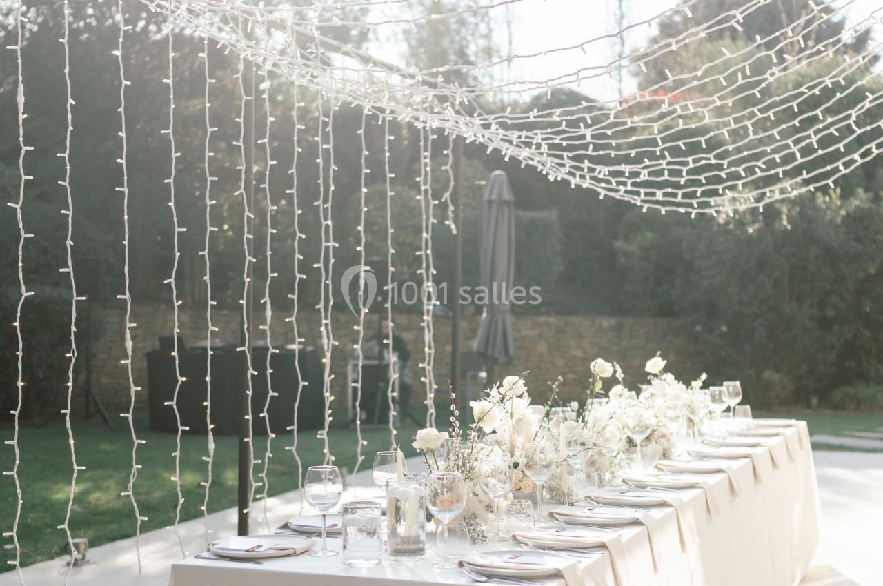 Table élégamment dressée en extérieur, décorée de fleurs blanches et éclairée par des guirlandes lumineuses suspendues.